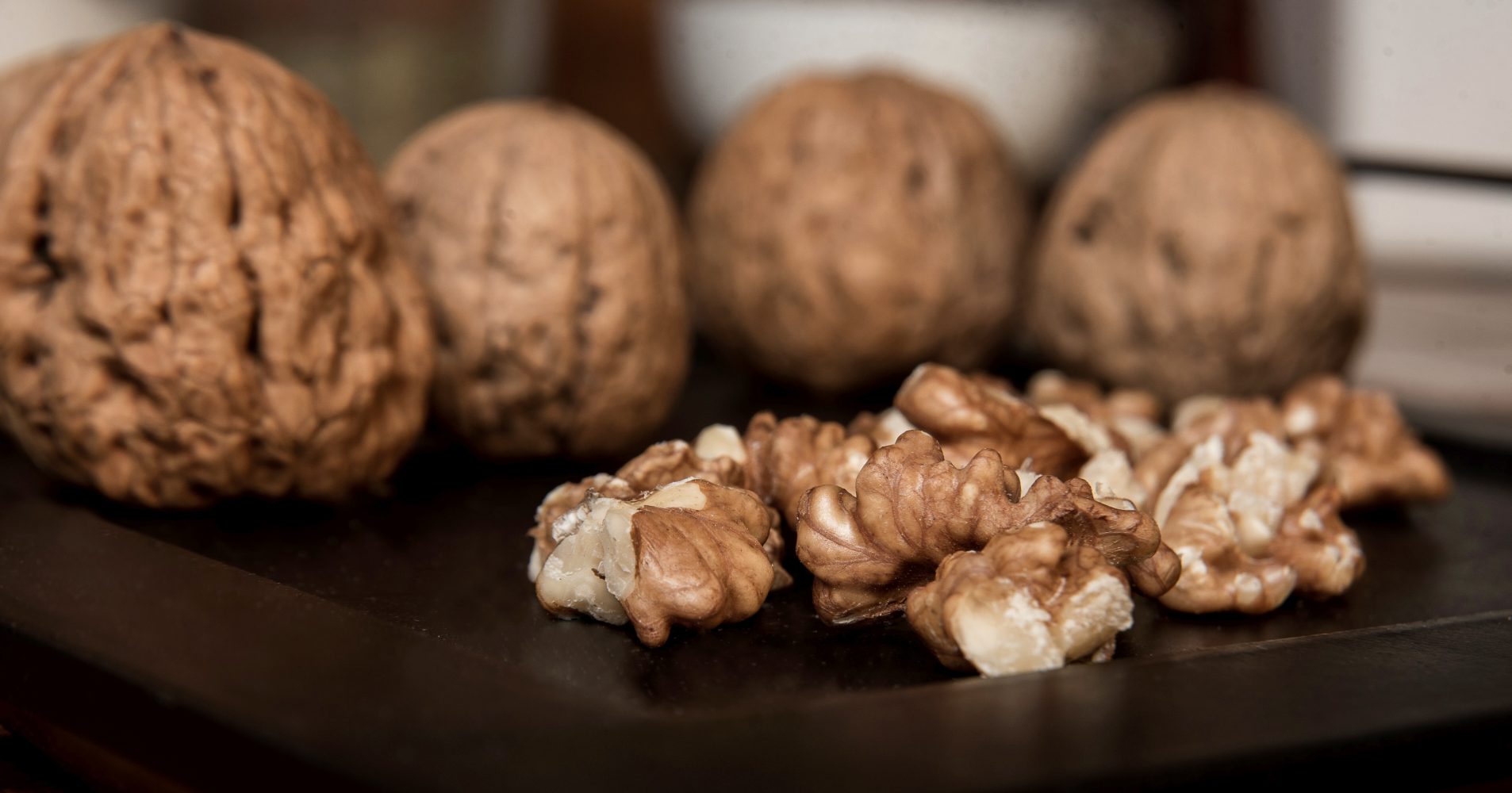 Primo piano di gherigli di noce sbucciati su una superficie scura, con diverse noci intere nel guscio sfocate sullo sfondo - Close-up of shelled walnut kernels on a dark surface, with several whole walnuts in their shells blurred in the background