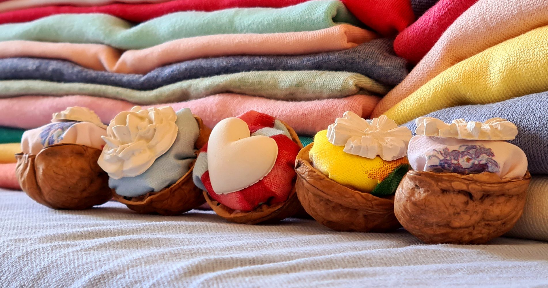 Cinque decorazioni artigianali realizzate con gusci di noce riempiti di stoffe colorate e piccoli gessetti a forma di fiore e cuore, con pile di maglioni colorati sullo sfondo - Five handmade decorations made from walnut shells filled with colorful fabrics and small chalk shapes in the form of flowers and hearts, with piles of colorful sweaters in the background