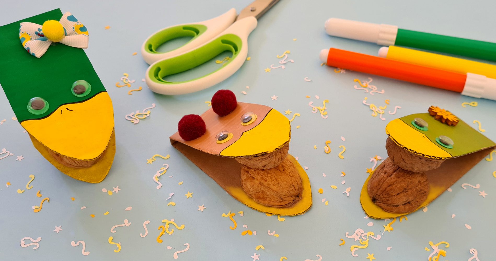 Tre paperelle realizzate a mano con cartoncino colorato e gusci di noce, disposte su un tavolo azzurro insieme a forbici e pennarelli - Three handmade ducklings made from colored cardboard and walnut shells, arranged on a light blue table with scissors and markers