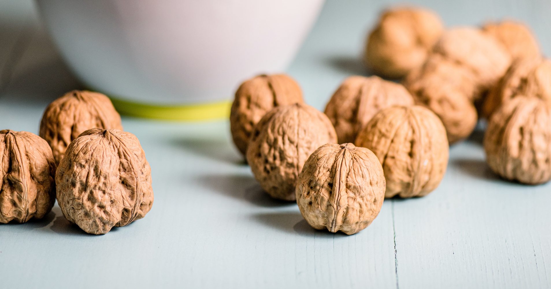 Diverse noci intere con guscio sparse su una superficie di legno azzurro chiaro, con una ciotola sfocata sullo sfondo - Several whole walnuts in their shells scattered across a light blue wooden surface, with a blurred bowl in the background