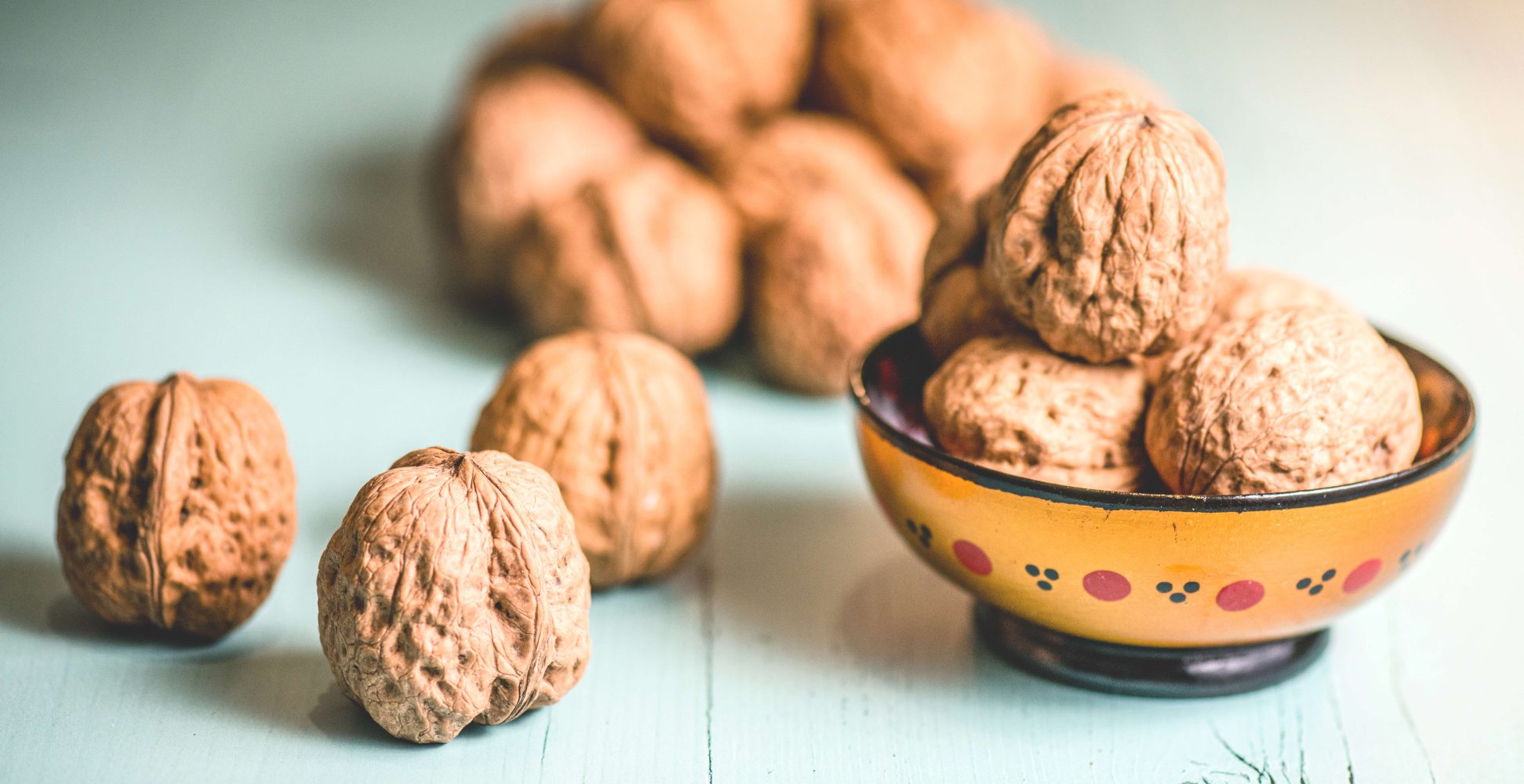 Noci intere con guscio disposte su un tavolo di legno azzurro, con alcune contenute in una piccola ciotola decorata in primo piano - Whole walnuts in their shells arranged on a light blue wooden table, with some placed in a small decorated bowl in the foreground