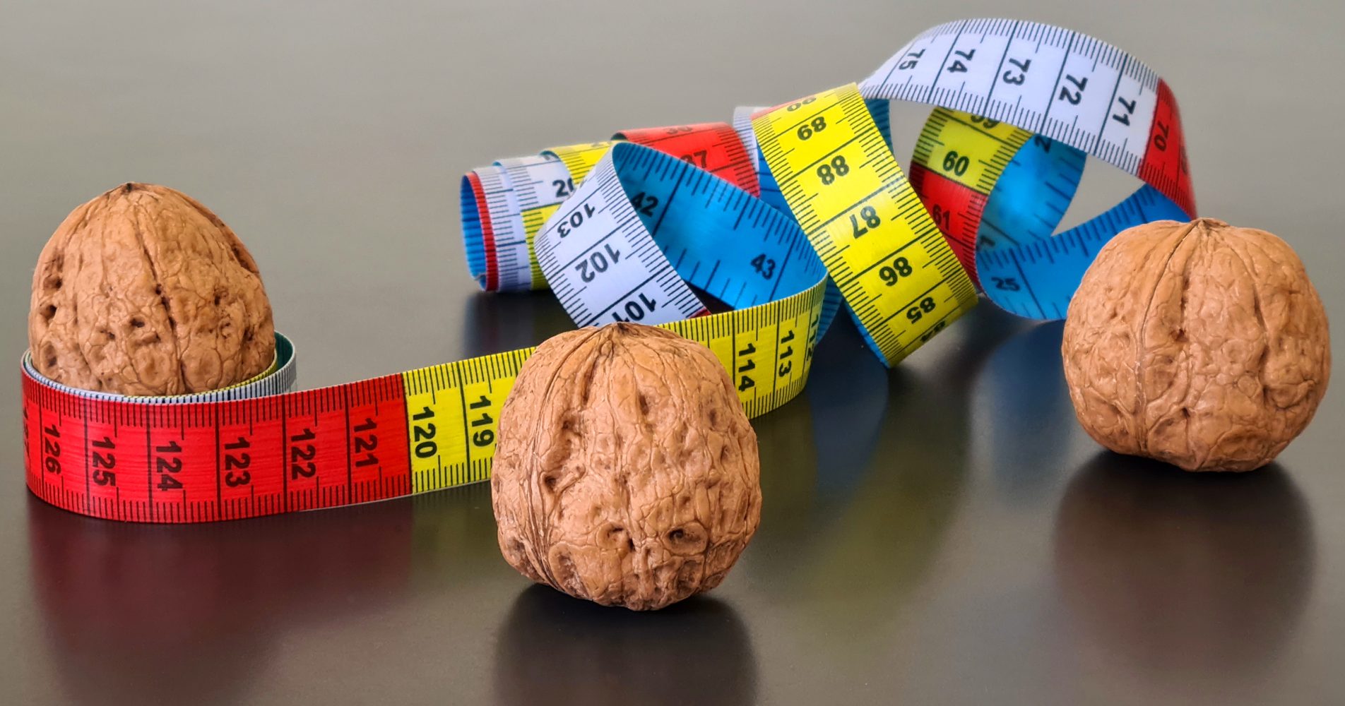 Tre noci intere disposte su un piano scuro accanto a un metro da sarta colorato - Three whole walnuts arranged on a dark surface next to a colorful measuring tape