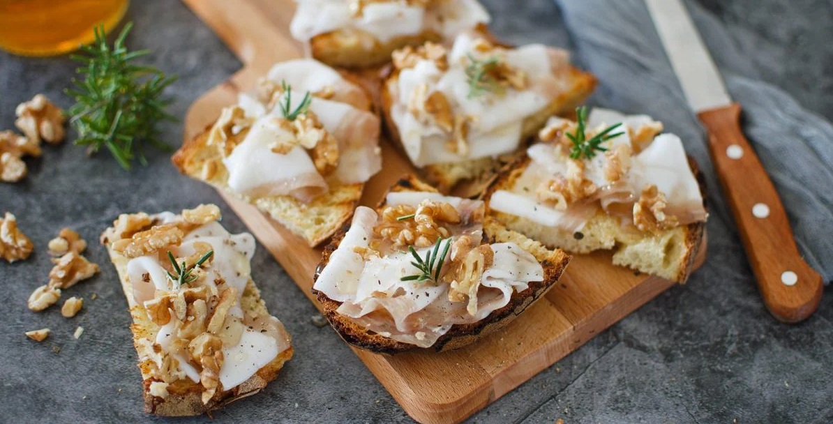 Bruschette di pane rustico tostato su un tagliere di legno, condite con sottili fette di lardo, gherigli di noce sbriciolati, un filo di miele e aghetti di rosmarino fresco - Toasted rustic bread bruschetta on a wooden cutting board, topped with thin slices of lard, crumbled walnut kernels, a drizzle of honey, and fresh rosemary needles