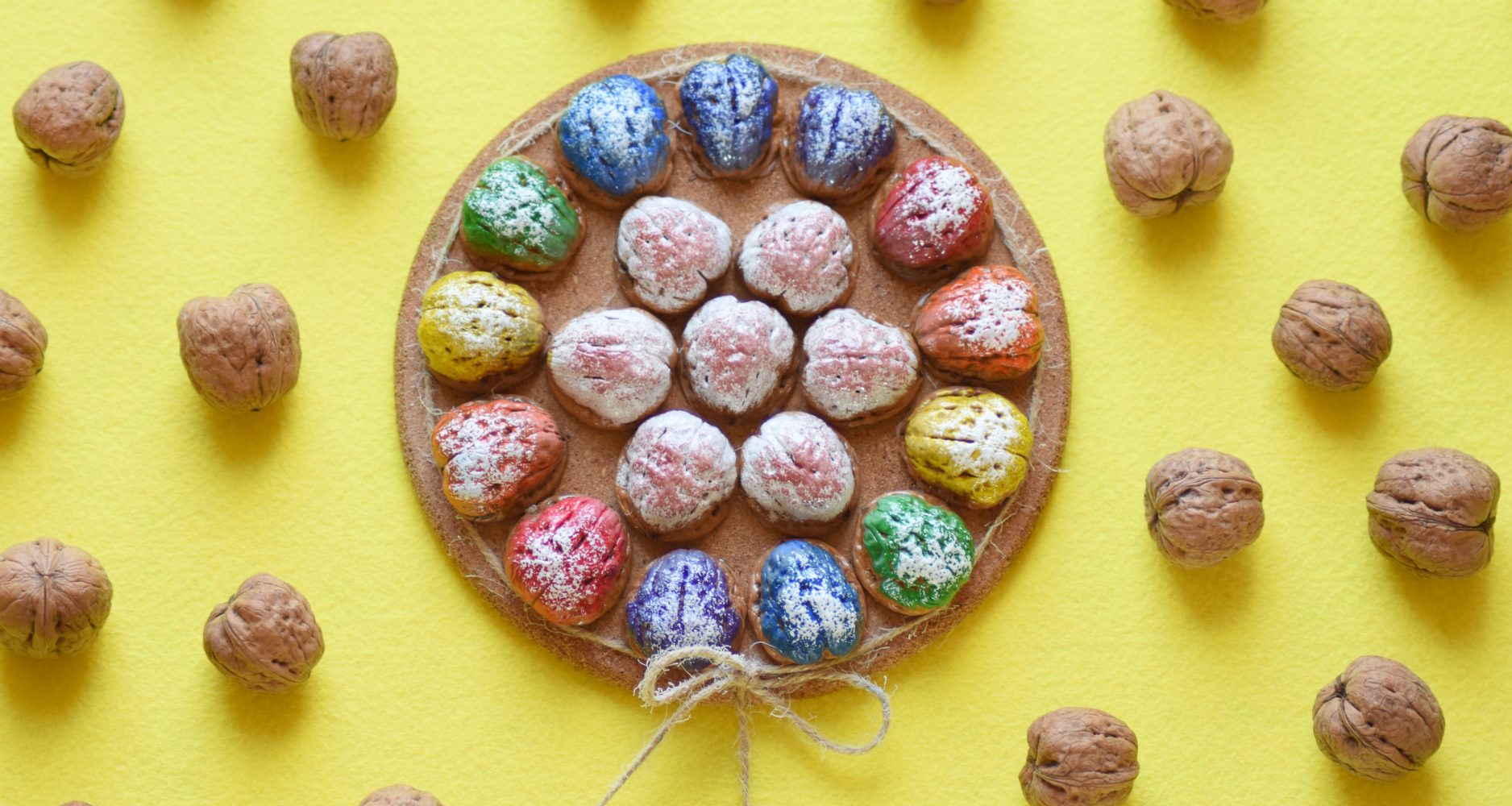piatto di sughero centrale decorato con noci dipinte in vari colori, circondato da noci naturali sparse su uno sfondo giallo - Central cork plate decorated with walnuts painted in various colors, surrounded by natural walnuts scattered on a yellow background