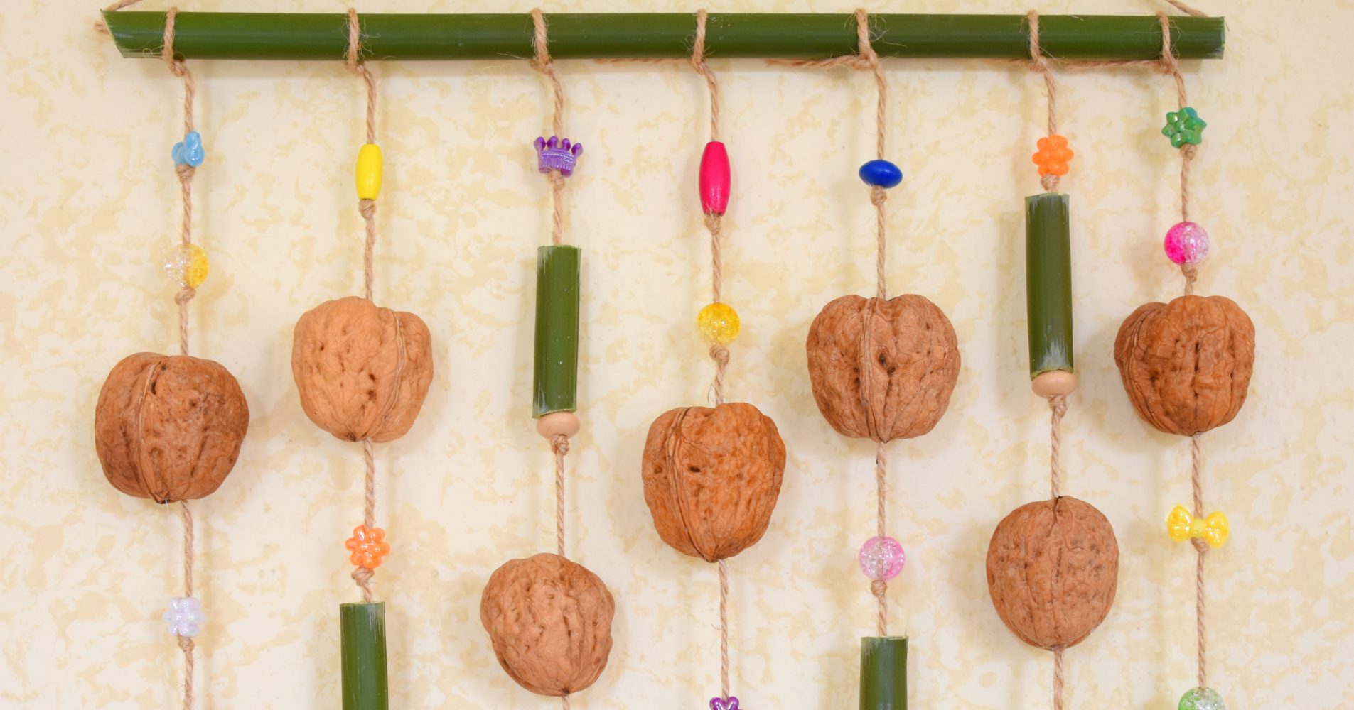 Decorazione da parete fatta a mano con noci intere, segmenti di bambù e perline colorate appesi a un bastoncino di bambù con spago - Handmade wall decoration with whole walnuts, bamboo segments, and colorful beads hanging from a bamboo stick with twine
