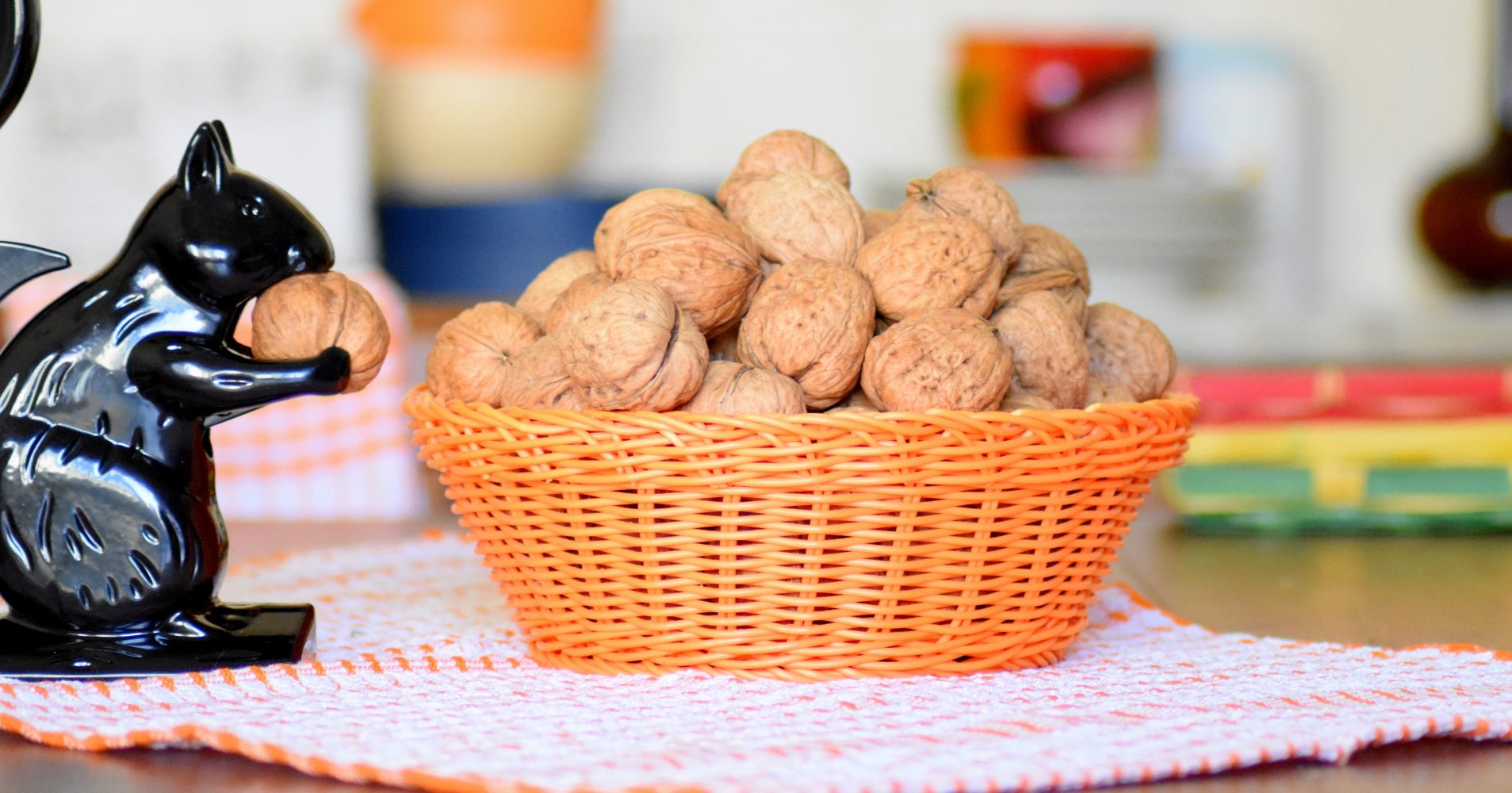 Un piccolo cesto arancione intrecciato pieno di noci con il guscio, posizionato su un tappetino a quadretti, accanto a un piccolo scoiattolo nero di plastica che tiene una noce tra le zampe - A small woven orange basket filled with whole walnuts, placed on a checkered mat, next to a small black plastic squirrel holding a walnut in its paws.