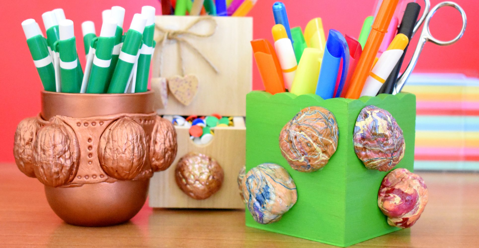 Tre portapenne artigianali decorati con gusci di noci su una scrivania in legno, con penne colorate e uno sfondo rosso vivace - Three handmade pen holders decorated with walnut shells on a wooden desk, with colorful pens and a bright red background