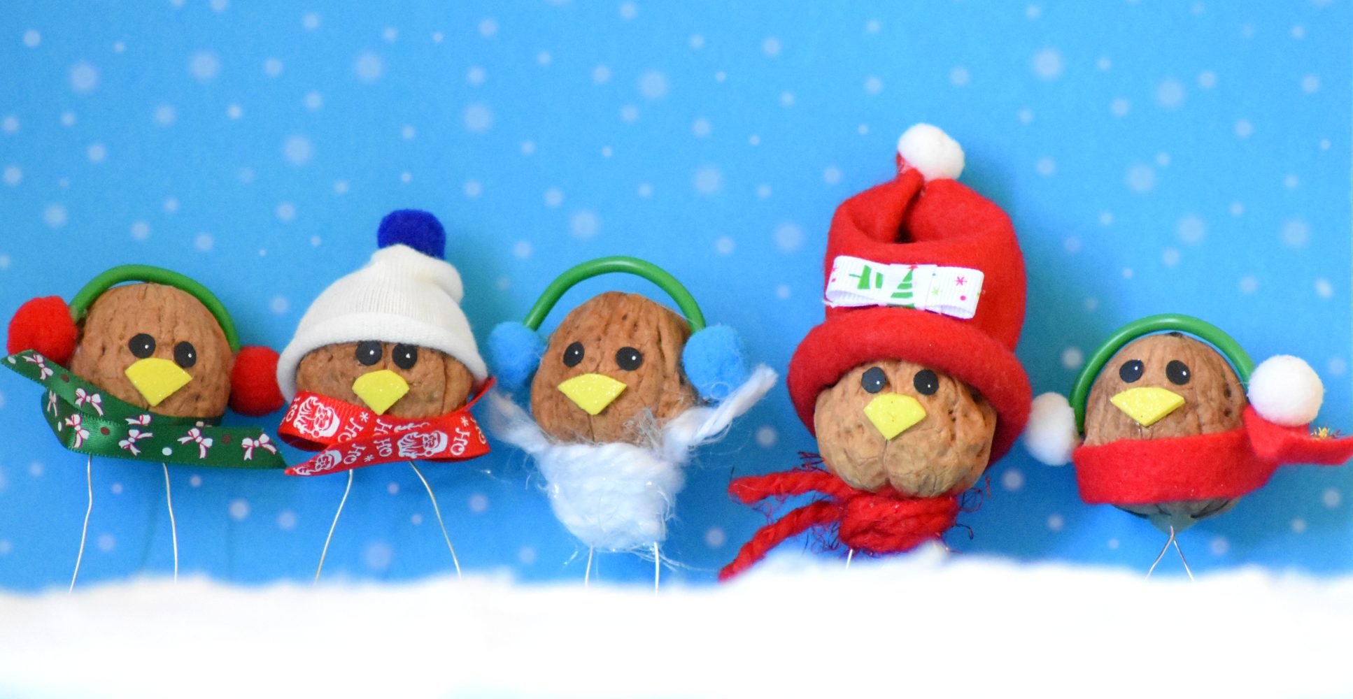 Cinque noci decorate come piccoli uccellini invernali con cappellini, sciarpe e paraorecchie colorati, disposte in fila su finta neve contro uno sfondo azzurro a pois bianchi che simula una nevicata - Five walnuts decorated as small winter birds wearing colorful hats, scarves, and earmuffs, arranged in a row on artificial snow against a light blue background with white polka dots simulating snowfall