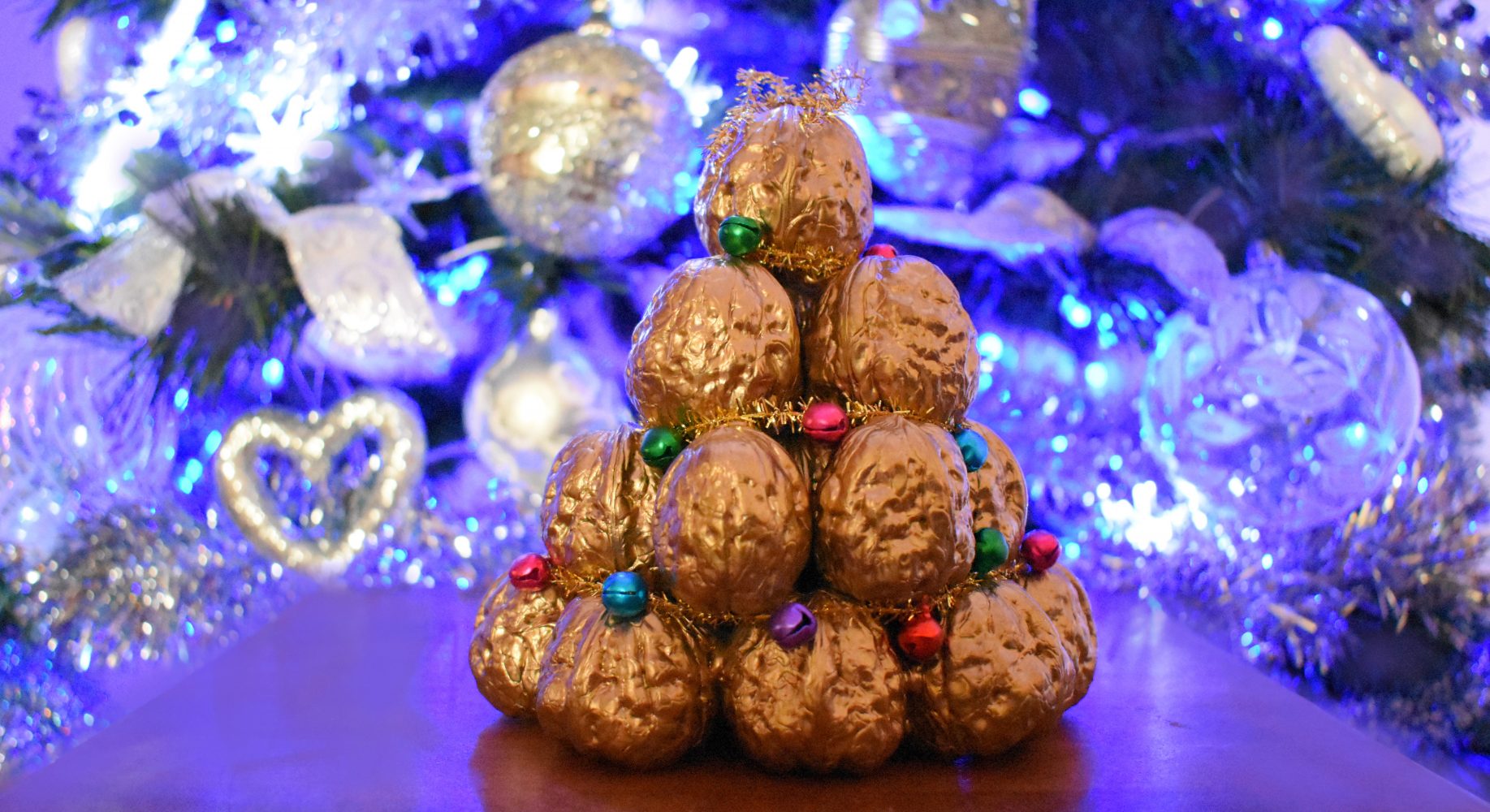 Piccolo albero di Natale a forma di piramide realizzato con noci dipinte d'oro, decorato con campanellini colorati. Sullo sfondo, un albero di Natale con luci blu sfocate - Small Christmas tree in the shape of a pyramid made of gold-painted walnuts, decorated with colorful bells. In the background, a Christmas tree with blurred blue lights