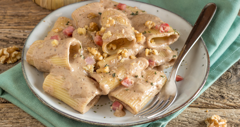 Un piatto di pasta tipo paccheri conditi con una densa crema di noci, guarniti con gherigli di noce tritati e cubetti di pancetta rosolata, serviti su un piatto bianco con bordo scuro - A plate of paccheri pasta coated in a rich walnut cream sauce, garnished with chopped walnut kernels and crispy browned pancetta cubes, served on a white plate with a dark rim