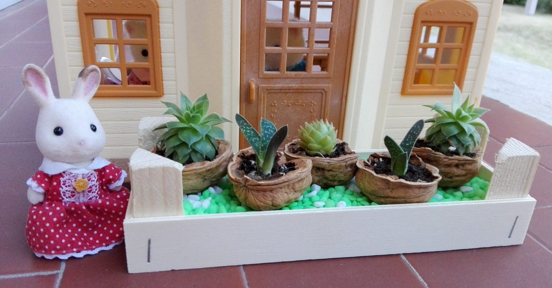 coniglietta accanto a una fioriera contenente piante davanti a una casa giocattolo | A little rabbit next to a planter with plants in front of a toy house