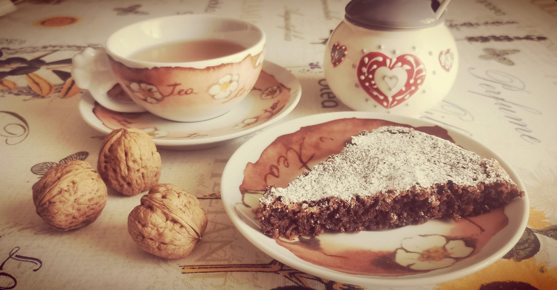Una fetta di torta al cioccolato spolverata di zucchero a velo su un piattino decorato, accanto a una tazza di tè fumante e tre noci intere - A slice of chocolate cake dusted with powdered sugar on a decorated saucer, next to a steaming cup of tea and three whole walnuts