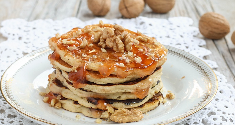 Una pila di cinque pancake soffici su un piatto bianco decorato, guarniti con abbondante sciroppo ambrato e gherigli di noce. Sullo sfondo, alcune noci intere su un tavolo di legno con un centrino di pizzo bianco - A stack of five fluffy pancakes on a decorated white plate, topped with plenty of amber syrup and walnut kernels. In the background, some whole walnuts rest on a wooden table with a white lace doily