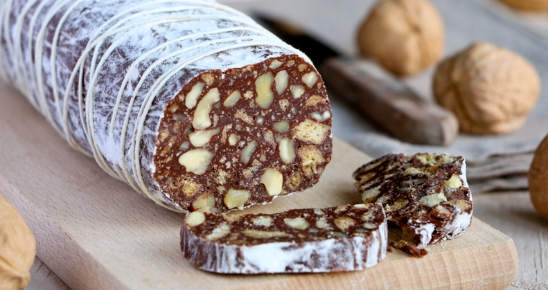Salame di cioccolato alle noci affettato su un tagliere di legno, con l'interno che mostra pezzi di noci e biscotti, circondato da noci intere con guscio sullo sfondo - Chocolate salami with walnuts, sliced on a wooden cutting board, showing an interior with walnut pieces and biscuits, surrounded by whole walnuts in their shells in the background