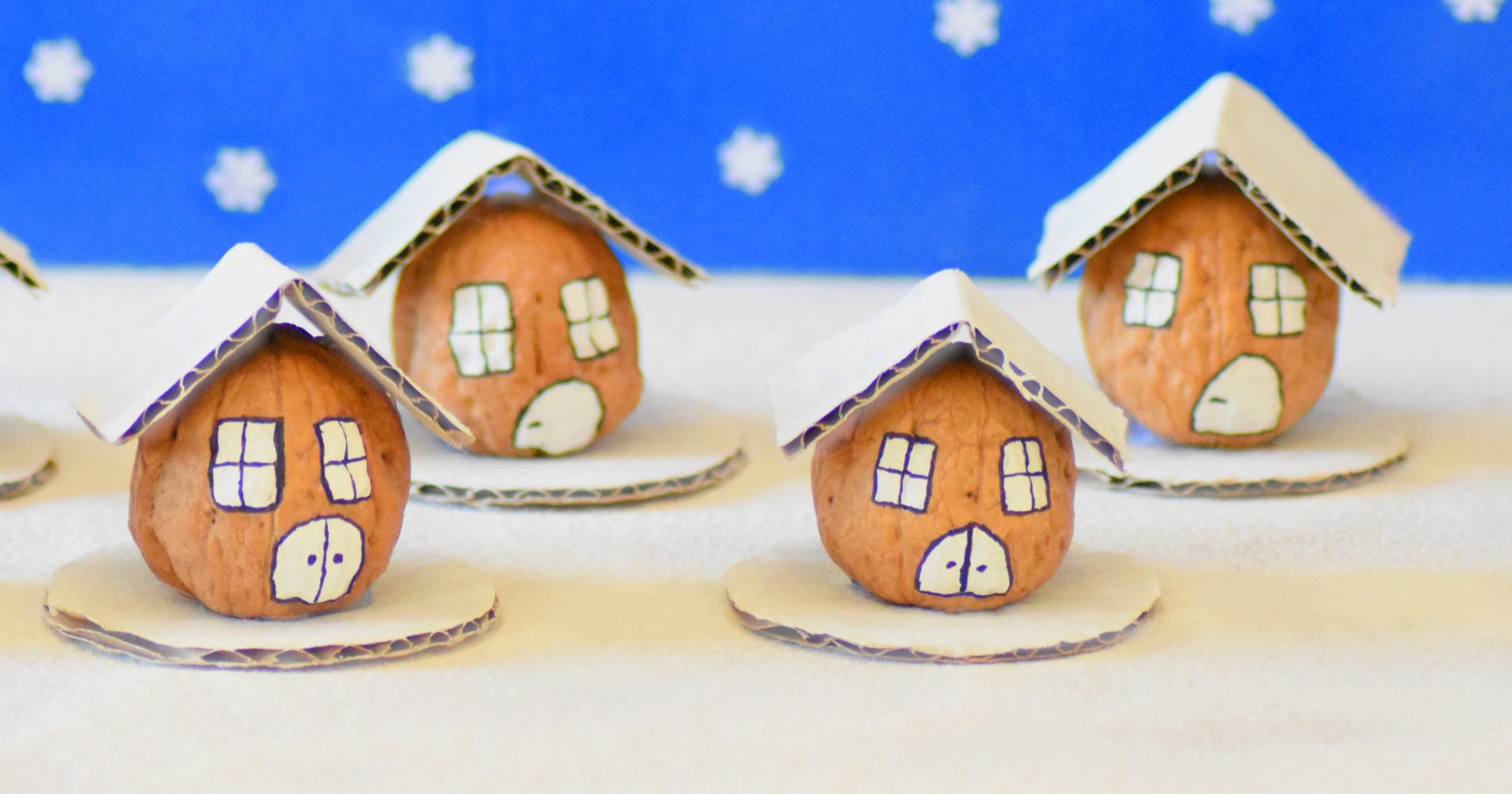 Quattro piccole casette natalizie realizzate con gusci di noce, decorate con finestre disegnate e tetti in cartoncino bianco, disposte su un piano bianco davanti a uno sfondo blu con fiocchi di neve - Four small Christmas houses made from walnut shells, decorated with drawn windows and white cardboard roofs, arranged on a white surface in front of a blue background with snowflakes