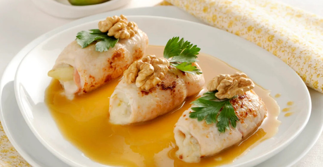 Tre involtini di carne dorati serviti su un letto di salsa bruna, guarniti con gherigli di noce e foglie di prezzemolo fresco, presentati su un piatto bianco - Three golden-brown meat rolls served on a bed of brown sauce, garnished with walnut kernels and fresh parsley leaves, presented on a white plate