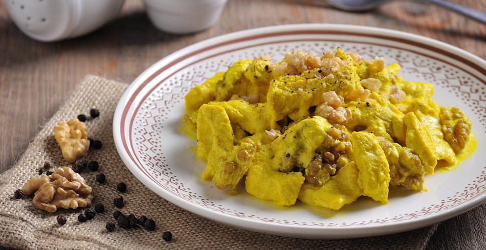 Bocconcini di pollo alla curcuma e yogurt guarniti con gherigli di noci, serviti su un piatto decorato accanto a grani di pepe nero e noci intere - Turmeric and yogurt chicken bites garnished with walnut kernels, served on a plate alongside black peppercorns and whole walnuts